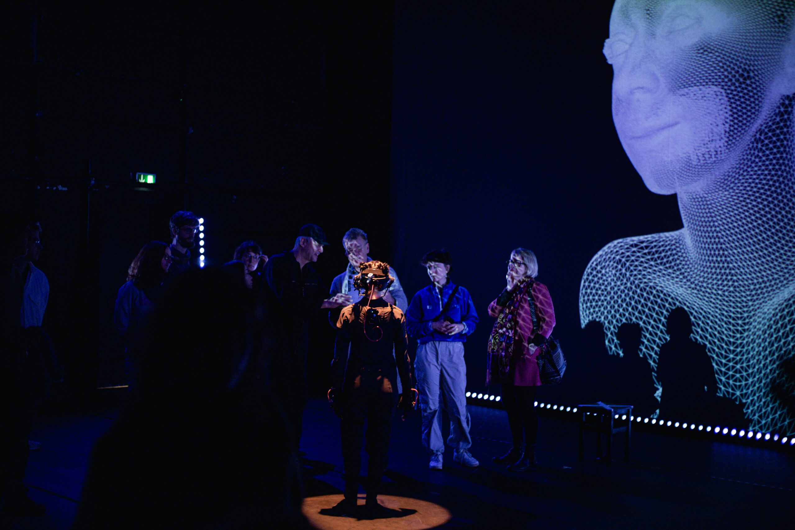 Performer in a motion capture suit with a spotlight surrounded by around 7 people, one explaining to the others. Projection in the background shows a mesh of an avatar