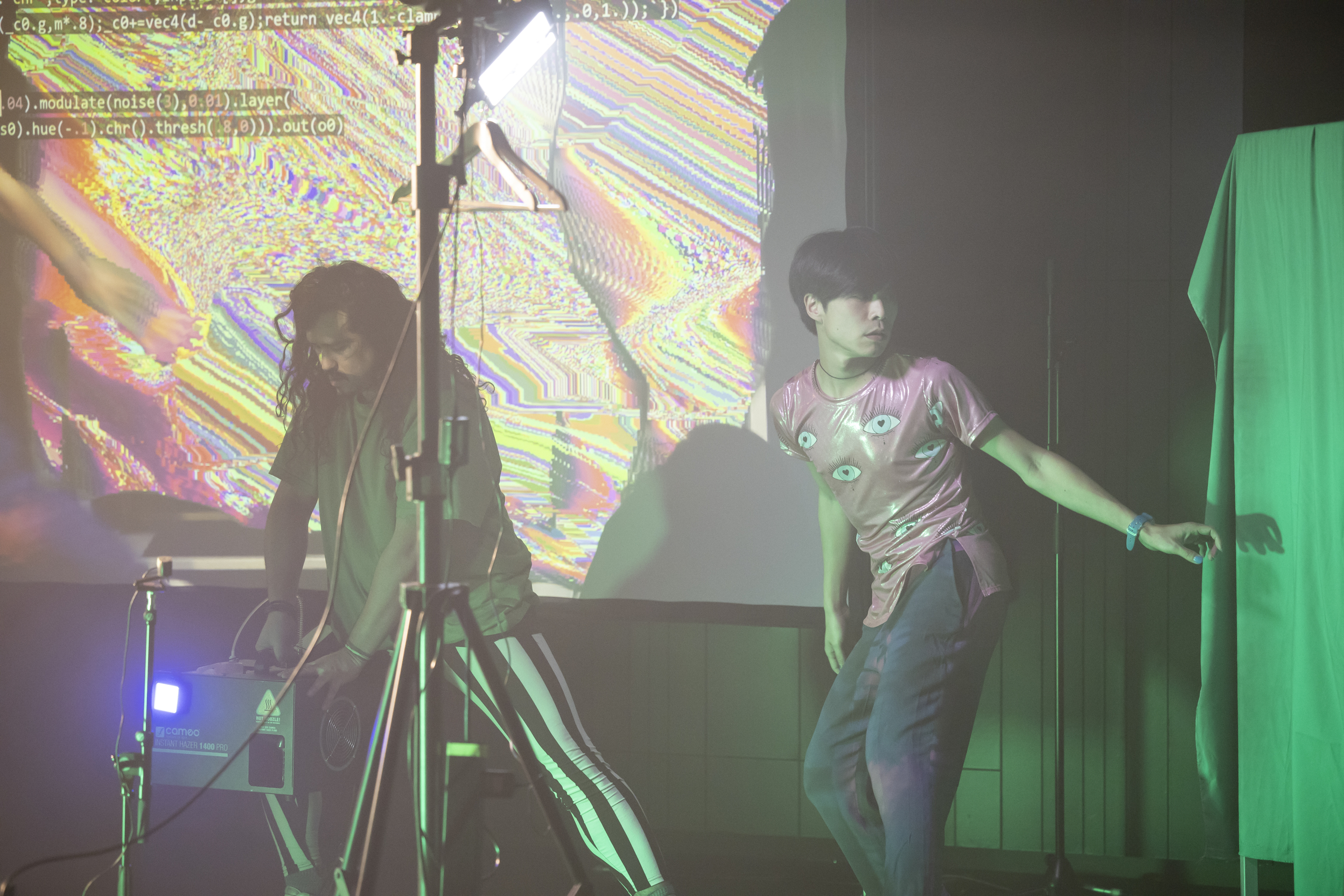 Two performers on stage facing away from each other, one holding a cubic machine and standing in front of glitchy projection