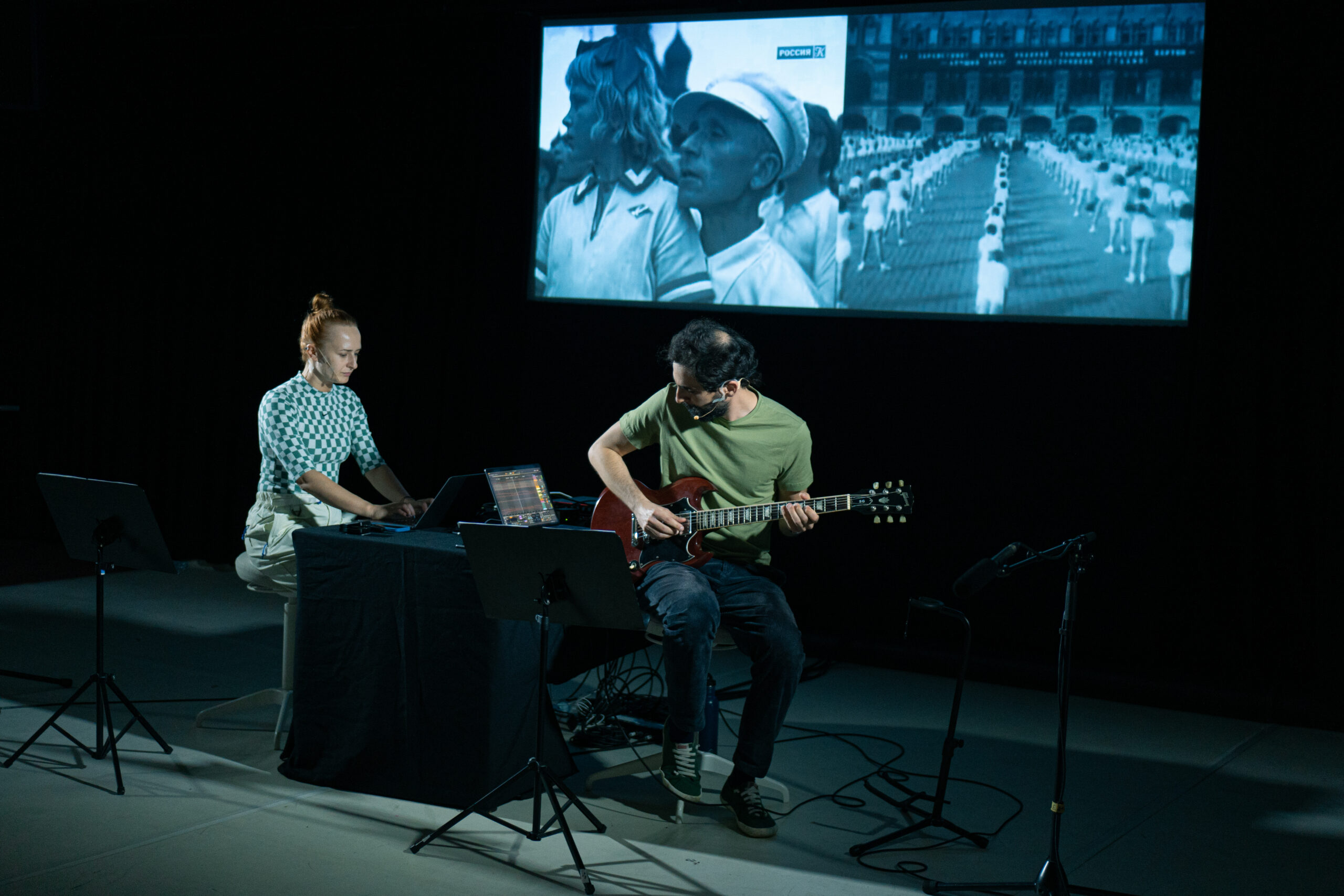 Two performers on stage, one operating a laptop and the other playing a guitar looking at a score. Projection in the background shows footage of a mass dance